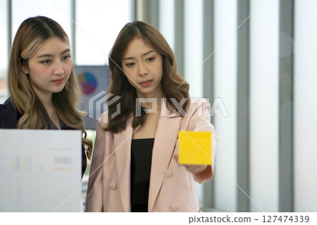 A professional woman stand together, engaged in a brainstorming session, using document and colorful paper notes on a transparent glass wall. 127474339