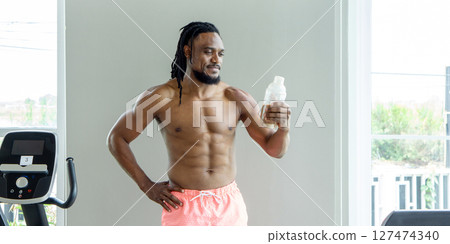 A muscular man stand shirtless, looking at protein shake bottle in his hand. He appear focused and dedicated to his fitness regimen. The atmosphere after an intense workout session. 127474340