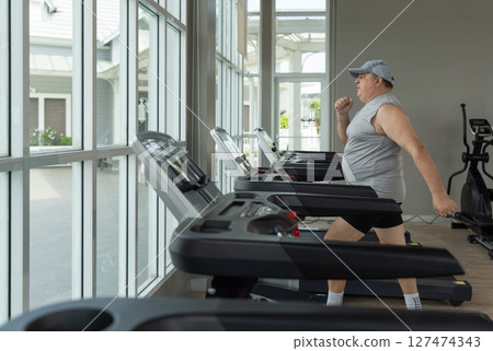 An older fat man walk with swinging arms on treadmill in bright gym during daytime promoting fitness and health. 127474343