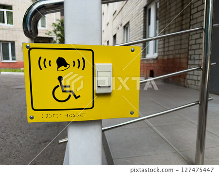 Modern inclusive call system for people with disabilities. Yellow panel with bell icon and wheelchair symbol fixed near accessible ramp outside residential building with tactile Braille marks 127475447