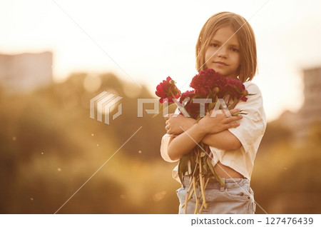 Holding beautiful red flowers. Little girl is on the summer field at daytime 127476439