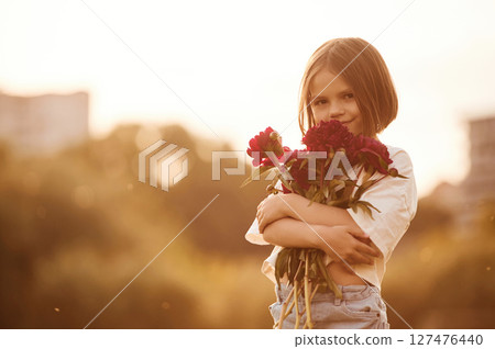 Holding beautiful red flowers. Little girl is on the summer field at daytime 127476440