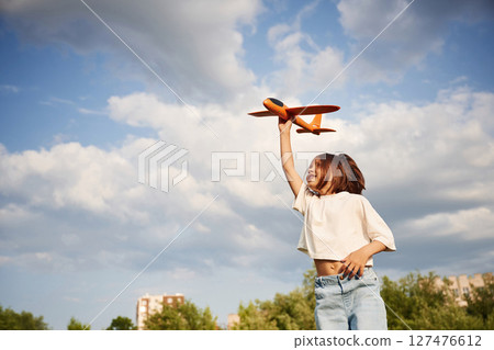Cloudy sky. Happy little girl is playing with toy plane outdoors Cloudy sky. Happy little girl is playing with toy plane outdoors 127476612