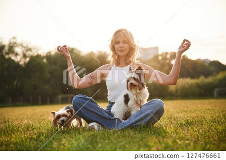 Sitting in yoga lotus pose. Young woman is with two little dogs on the summer field Sitting in yoga lotus pose. Young woman is with two little dogs on the summer field 127476661