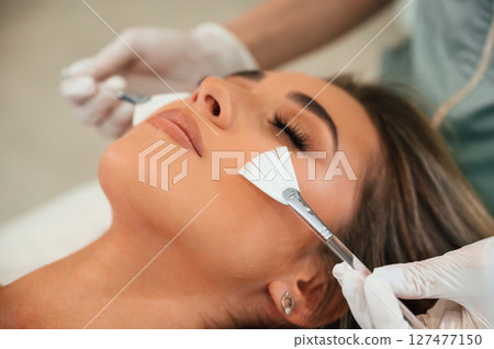 Cleaning skin by brushes. Woman face getting facial care by beautician hands at spa salon Cleaning skin by brushes. Woman face getting facial care by beautician hands at spa salon 127477150