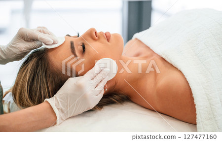 Cleaning skin by using cotton pad or sponge. Woman face getting facial care by beautician hands at spa salon 127477160