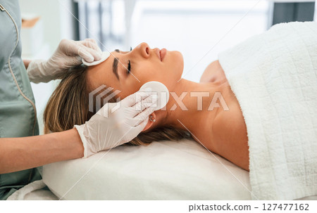 Cleaning skin by using cotton pad or sponge. Woman face getting facial care by beautician hands at spa salon Cleaning skin by using cotton pad or sponge. Woman face getting facial care by beautician hands at spa salon 127477162