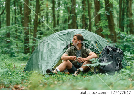 With cup of water. Sitting near the tent. Tourist in summer forest. Conception of exploration and leisure With cup of water. Sitting near the tent. Tourist in summer forest. Conception of exploration and leisure 127477647