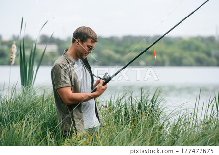 Standing near the lake. Handsome man in sunglasses is fishing outdoors 127477675