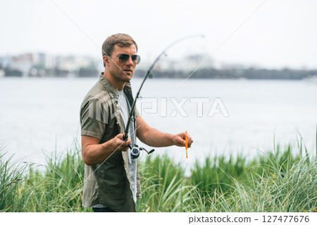 Standing near the lake. Handsome man in sunglasses is fishing outdoors 127477676