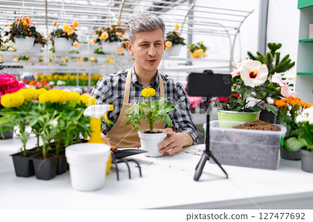 A man in a greenhouse demonstrates gardening techniques, holding a potted plant while recording a video. 127477692