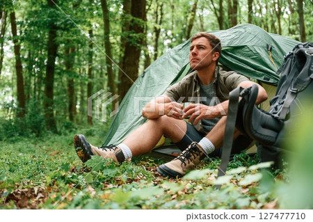 With cup of water. Sitting near the tent. Tourist in summer forest. Conception of exploration and leisure 127477710
