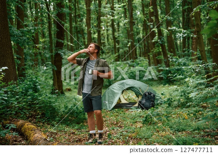 Green colored tent is behind. Tourist in summer forest. Conception of exploration and leisure Green colored tent is behind. Tourist in summer forest. Conception of exploration and leisure 127477711