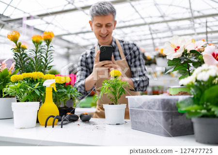 A smiling man in a plant nursery photographs a flower. Various plants, gardening tools, and pots fill the foreground. 127477725