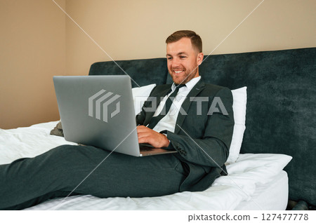 Positive businessman is indoors in the hotel room using laptop 127477778