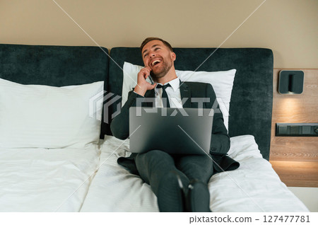 Smiling, lying down on bed and talking by phone. Businessman is indoors in the hotel room Smiling, lying down on bed and talking by phone. Businessman is indoors in the hotel room 127477781