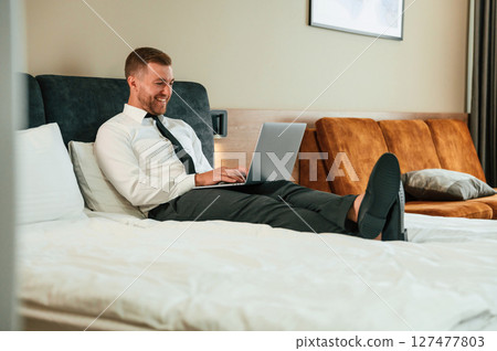 Working by lying down on the bed with laptop. Businessman is indoors in the hotel room Working by lying down on the bed with laptop. Businessman is indoors in the hotel room 127477803