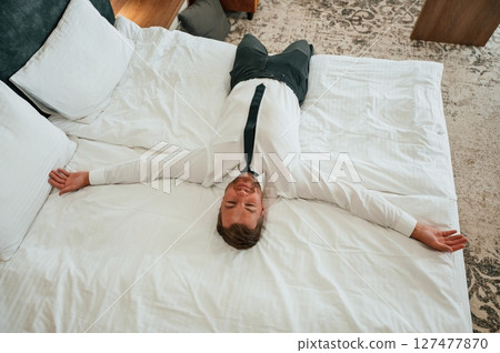 Tired, lying down on the bed. Businessman is indoors in the hotel room Tired, lying down on the bed. Businessman is indoors in the hotel room 127477870