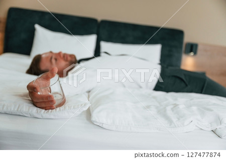 Empty glass in hand, lying down on the bed, tired. Businessman is indoors in the hotel room 127477874