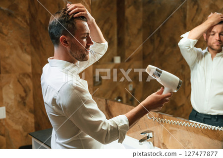 Drying his hair. Man in formal clothes is in the bathroom against mirror Drying his hair. Man in formal clothes is in the bathroom against mirror 127477877