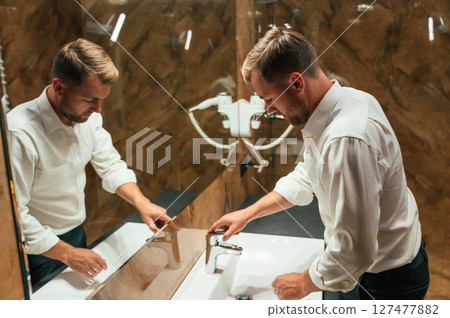 Washing hands. Man in formal clothes is in the bathroom against mirror Washing hands. Man in formal clothes is in the bathroom against mirror 127477882