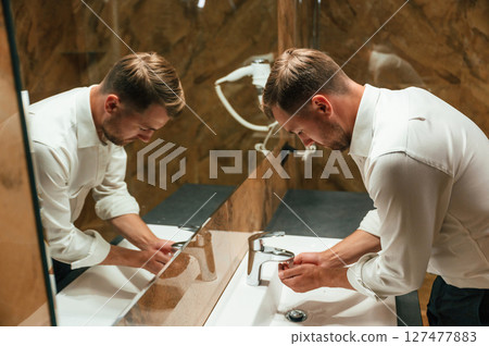 Washing hands. Man in formal clothes is in the bathroom against mirror Washing hands. Man in formal clothes is in the bathroom against mirror 127477883