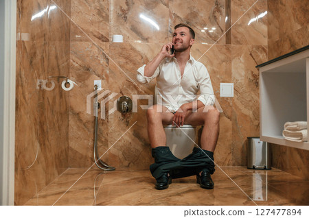 Having a call. A man in a white shirt is sitting on the toilet, holding a smartphone in his hands 127477894