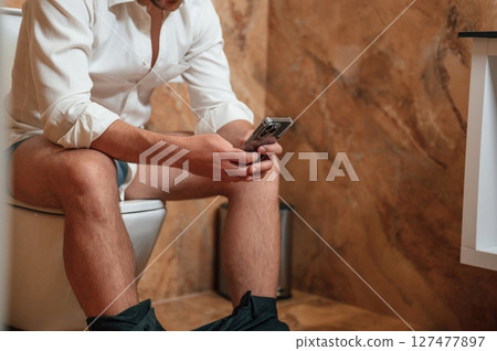 A man in a white shirt is sitting on the toilet, holding a smartphone in his hands 127477897