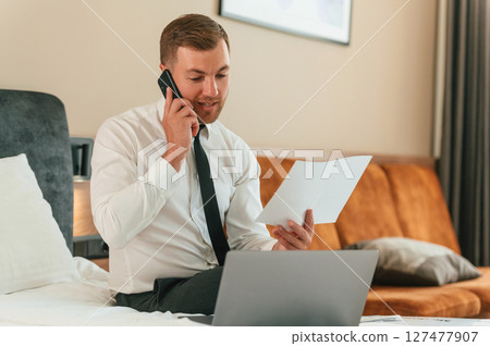 Reading the information from ticket, talking by phone. Businessman is indoors in the hotel room 127477907