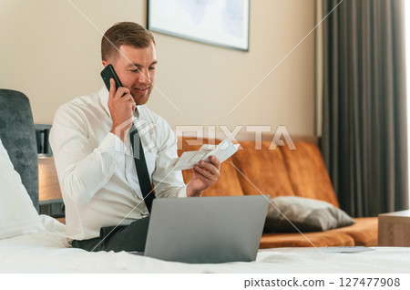 Reading the information from ticket, talking by phone. Businessman is indoors in the hotel room Reading the information from ticket, talking by phone. Businessman is indoors in the hotel room 127477908