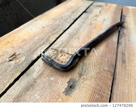 Close up view of steel pry bar placed diagonally on rough wooden workbench, captured in natural light, highlighting texture, wear marks and rustic workshop atmosphere Close up view of steel pry bar placed diagonally on rough wooden workbench, captured in natural light, highlighting texture, wear marks and rustic workshop atmosphere 127478298