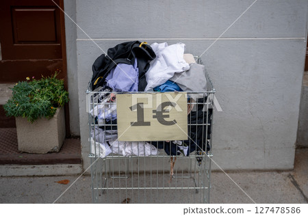 Used clothing in a metal crate with a 1 euro sign, placed outdoors, concept of thrift shopping Used clothing in a metal crate with a 1 euro sign, placed outdoors, concept of thrift shopping 127478586