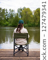 A woman sits on a portable chair by a calm river, enjoying the serene natural landscape 127479341