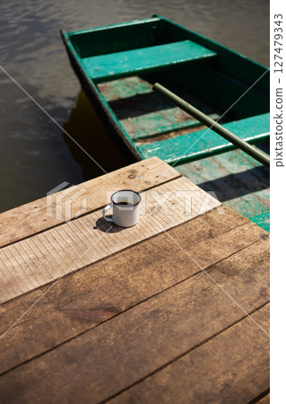 A rustic wooden dock by the water features a single enamel mug of coffee, offering a tranquil lakeside scene 127479343