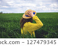 A woman in a yellow sweater and straw hat enjoys a sunny day in a vibrant green field 127479345