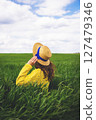 A woman in a yellow sweater and straw hat enjoys the tranquility of a green field, lost in contemplation under a partly cloudy sky 127479346