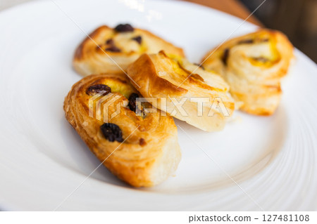 Three raisin pastries served on white plate during hotel breakfast. Morning dessert, sweet bakery snack, and elegant table setting concept. Three raisin pastries served on white plate during hotel breakfast. Morning dessert, sweet bakery snack, and elegant table setting concept. 127481108