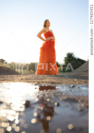 A Glamorous Model Dressed in a Stunning Orange Evening Gown Stands Beautifully at Sunset 127483451