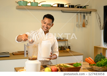 Smiling man adds protein powder to a blender while preparing a healthy smoothie 127483858