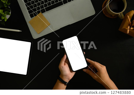 Hand holding smartphone with blank screen on dark desk workspace. Hand holding smartphone with blank screen on dark desk workspace. 127483880