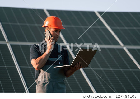 Talking by phone, holding laptop. Man is doing operating and maintenance in solar power plant Talking by phone, holding laptop. Man is doing operating and maintenance in solar power plant 127484103