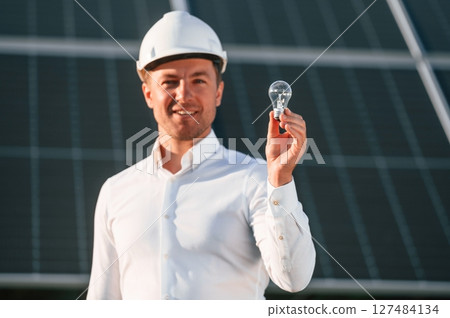 Man in white shirt and hart hat is holding bulb on a solar power plant Man in white shirt and hart hat is holding bulb on a solar power plant 127484134