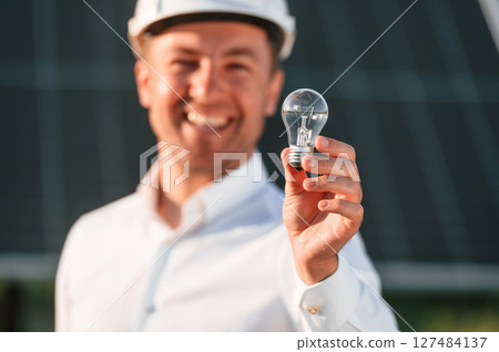 Man in white shirt and hart hat is holding bulb on a solar power plant Man in white shirt and hart hat is holding bulb on a solar power plant 127484137