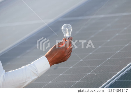 Conception of green energy. Close up view of man's hand that is holding light bult in front of solar panel Conception of green energy. Close up view of man's hand that is holding light bult in front of solar panel 127484147