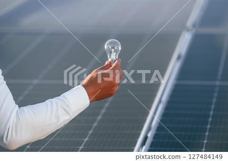 Close up view of man's hand that is holding light bult in front of solar panel 127484149