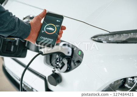 Using smartphone app for monitoring charging process. Close up view of man that is with electrical car Using smartphone app for monitoring charging process. Close up view of man that is with electrical car 127484421
