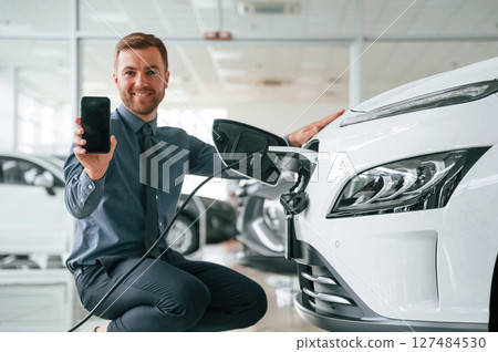 Sitting, showing smartphone, charging. Handsome car dealership worker is with electric vehicle Sitting, showing smartphone, charging. Handsome car dealership worker is with electric vehicle 127484530
