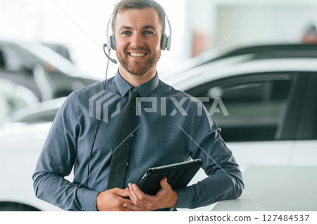 Manager is in the headphones and with notepad. Handsome car dealership worker is with electric vehicle 127484537