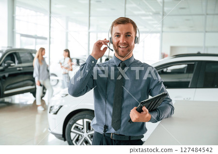 Manager is in the headphones and with notepad. Handsome car dealership worker is with electric vehicle 127484542
