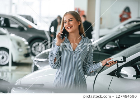 Talking by phone and holding charger. Beautiful woman is indoors with the modern car 127484590